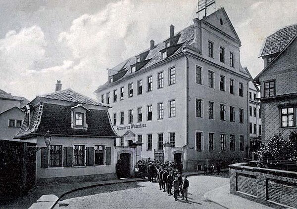 Schwarz-weiß Foto eines großen vierstöckigen Gebäudes mit der Aufschrift Reformiertes Waisenhaus. Das Gebäude liegt in einer gepflasterten Strasse, die als Rechtskurve abbiegt. Link im Bild ein kleines Pförtnerhaus und ein von Mauern gerahmtes eisernes Tor. Eine Gruppe Waisenjungen in Zweierreihe kommt gerade aus dem Tor.