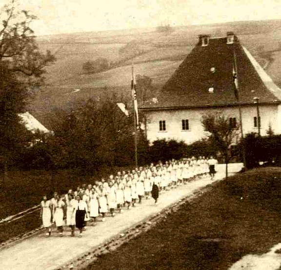 Viele weis gekleidete Mädchen marschieren in Dreierreihen auf einer Straße, daneben Erzieherinen 