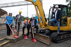 5 Personen mit Schaufeln und Bagger beim symbolischwn Spatenstich 2021