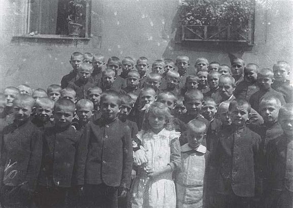 Schwarz-weiß Gruppenfoto mit Emilie Wilhelm als einziges Mädchen umgeben von 46 Waisenhausjungen und einem Erwachsenen.Sie hält eine Puppe im Arm und trägt ein weißes Kleid. Die Jungen stehen in vier Reihen und sind kurz geschoren. Alle sind dunkel uniform gekleidet, nur ein Junge neben Emilie ist ebenfalls hell mit weißem Kragen. Der Erwachsene trägt einen imposanten Schnurrbart 