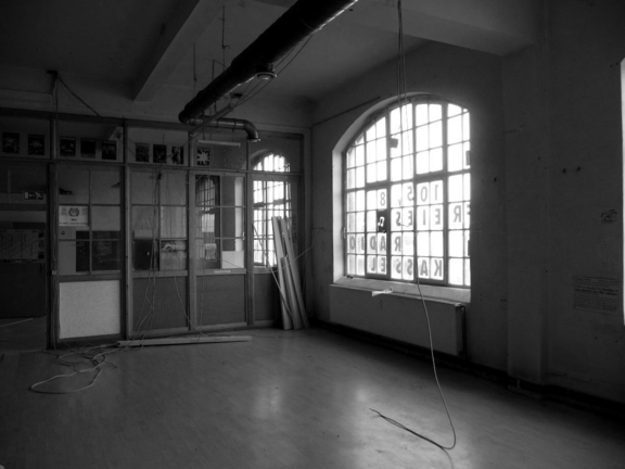 Schwarzweiß Foto eines leeren Raums in der Salzmann-Fabrik mit spiegelverkehrter Werbe-Beschriftung an den Fenstern. Von der Decke hängen Kabel, in einer Ecke stapeln sich gebrauchte Kabelschächte, ein Schild und Plakate kleben an einer gläsernen Trennwand. 