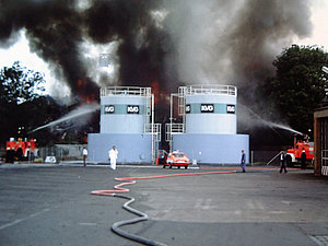 Zwei Löschfahrzeuge kühlen mit Wenderohren zwei Öllagertanks, dahinter Rauchpilz