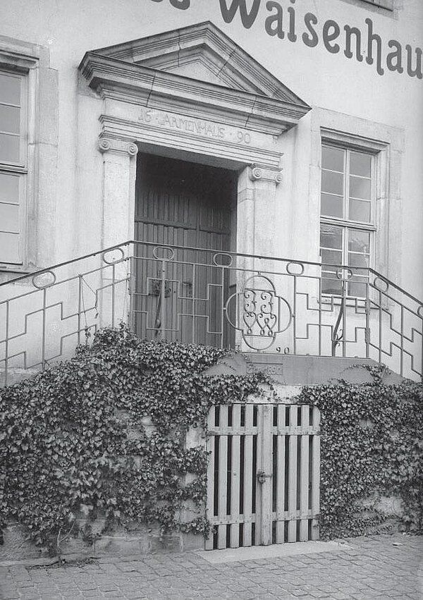 Schwarz-weiß Foto einer großen Eingangstür mit zweiseitiger Treppe. Die Tür ist von jonischen Pilastern und einem klassischen Dreiecksgiebel umrahmt. Die Freitreppe Geländer wird von Wein bewuchert. Mittig frontal ein holzvergittereter Keller unter der Treppe.
