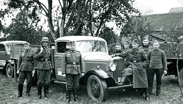 Fertig zur erzwungenen Übergabe an das Militär in einem Garten. Mehere Personen in Uniform, dritte Person von links Hans Hess.