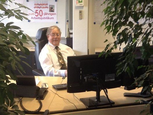 Alwin Krönert sitzt in seinem Büro am Schreibtisch, dahinter Plakat zu seinem 50jährigen Arbeitsjubiläum, 2015 