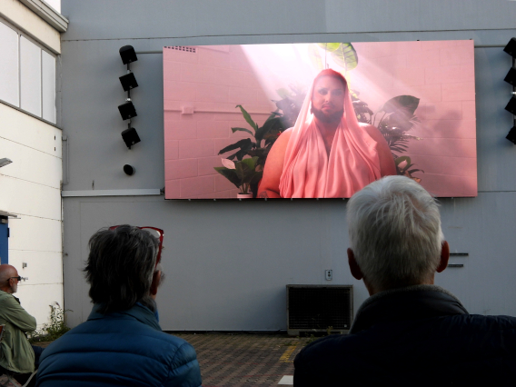 Zwei Besucher von hinten gesehen sitzen vor einer Videoleinwandauf auf der ein Mann im rosa Gewand zu sehen ist 